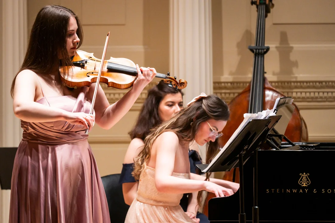 A pianist sits and plays a piano while a violinist stands next to her playing the violin.