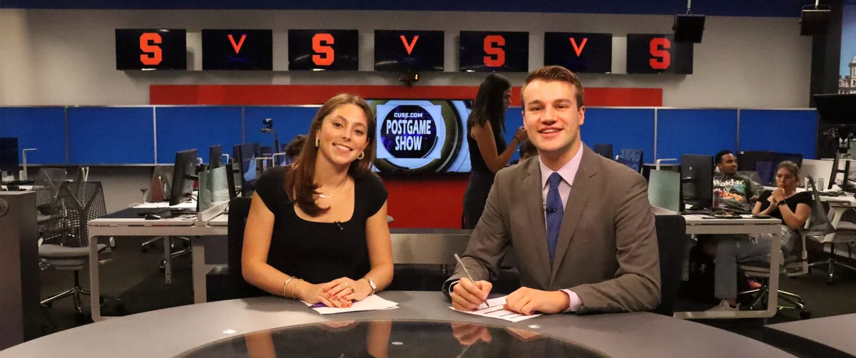 Two student journalists posing at an anchor table during a broadcast