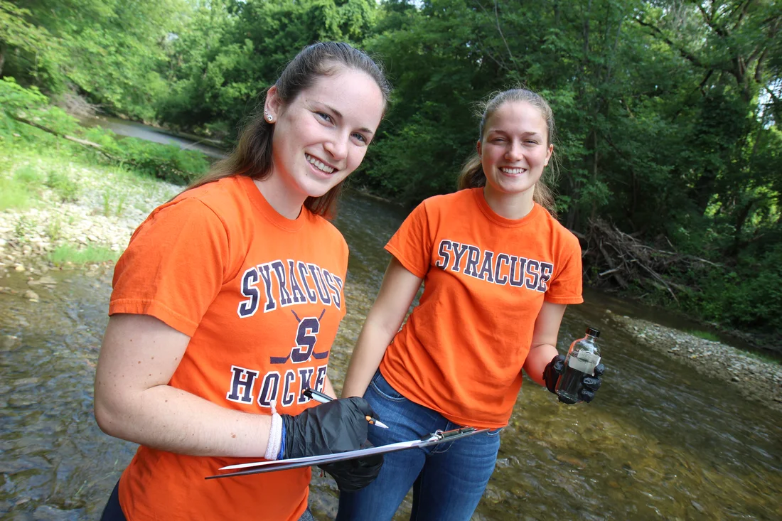 Two students standing in a stream, one holding water sample in a jar and the other is holding a clipboard.