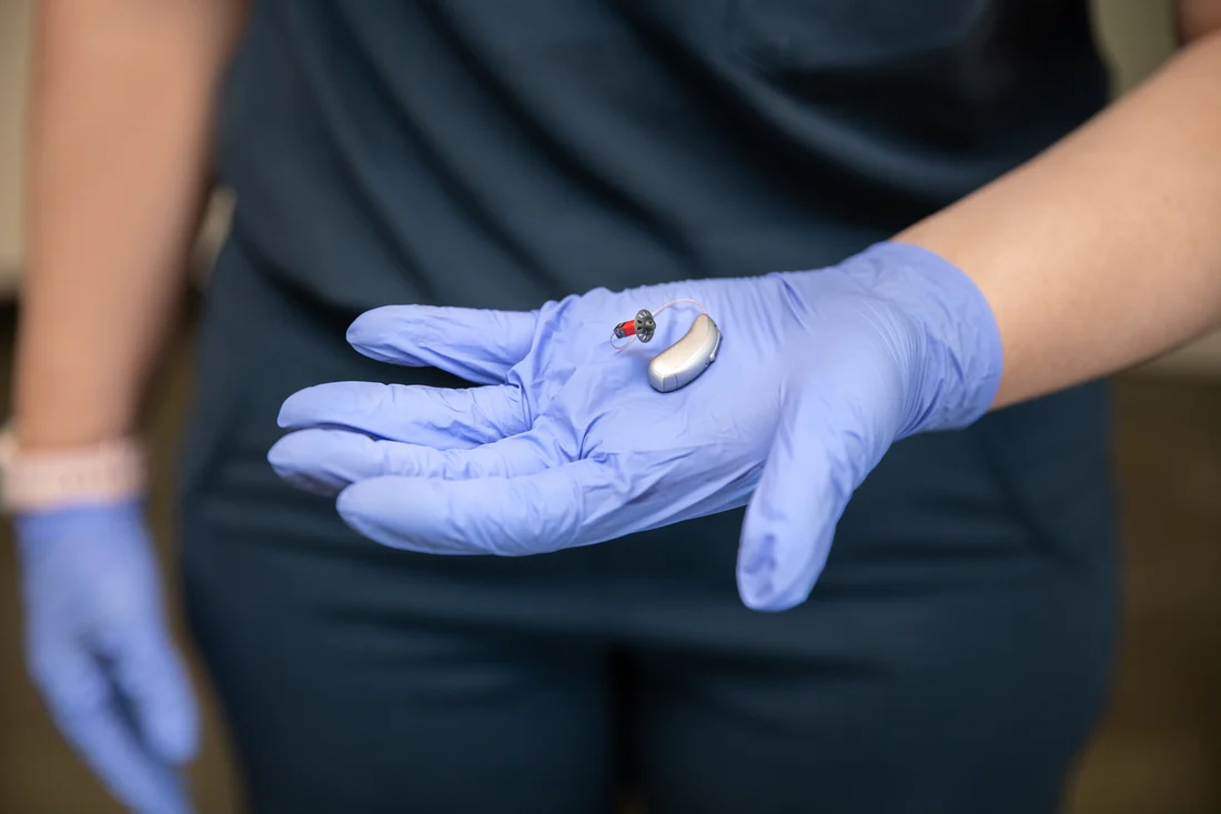 An image of a person's hand, wearing a nylon glove, holding a hearing aid
