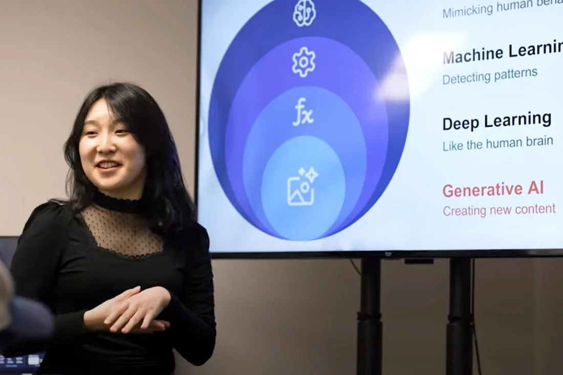 A student stands in front of a large screen, smiling while giving a presentation about AI.