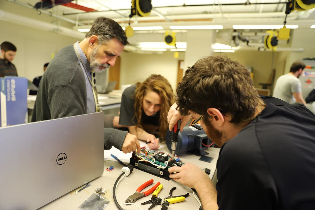 A professor points to a component while two students use tools to work on it