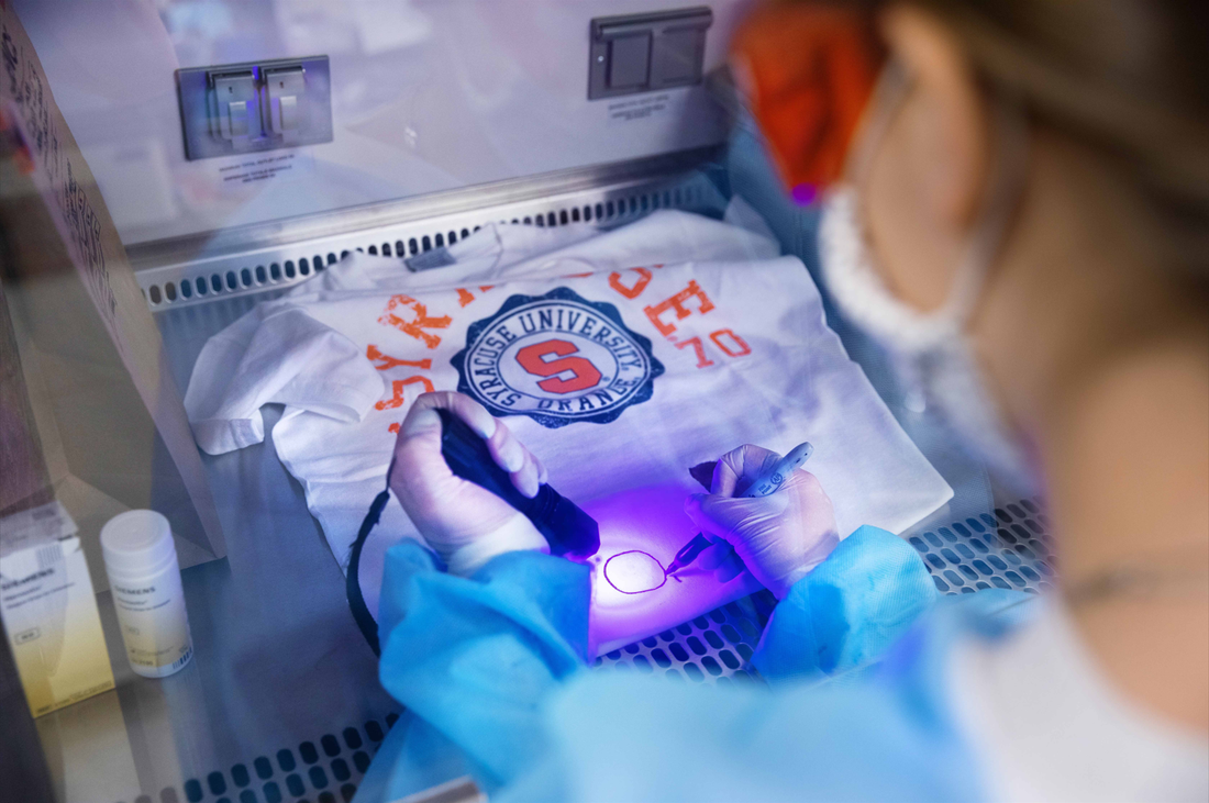 A student shines a UV light and creates a mark on a t-shirt, under a chemical hood in a forensics laboratory. The t-shirt is printed with an orange Syracuse logo.