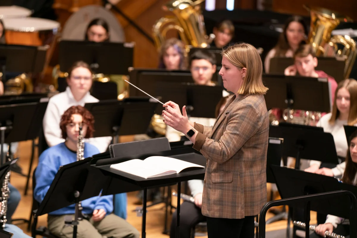 A person conducts a large ensemble of musicians in a historic auditorium.