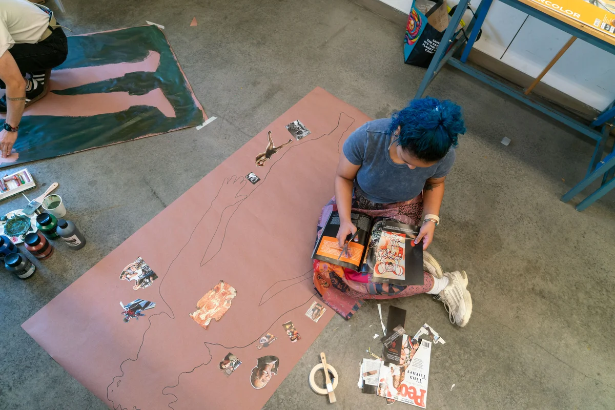 A person sits on the floor on a large piece of paper with the outline of a body drawn on it. The person is cutting out images from a magazine and laying them on different areas of the body outline.