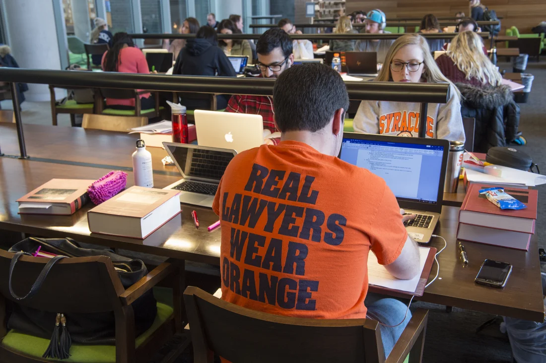 A student sitting at a desk in a campus library, facing away from the camera. The back of his shirt reads, "Real lawyers wear orange."